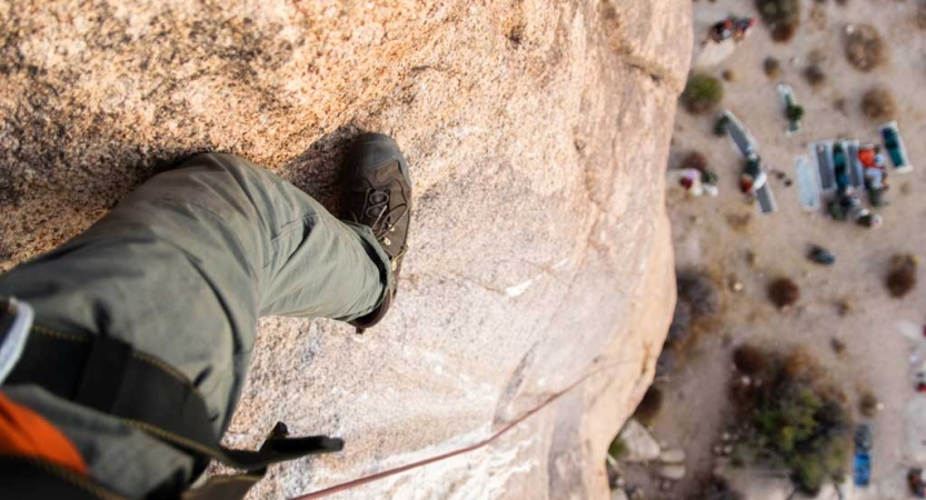 From the viewpoint of a rock climber looking down, you can see the safety gear rope attached to a leg. Far below are a group of people and their gear. 